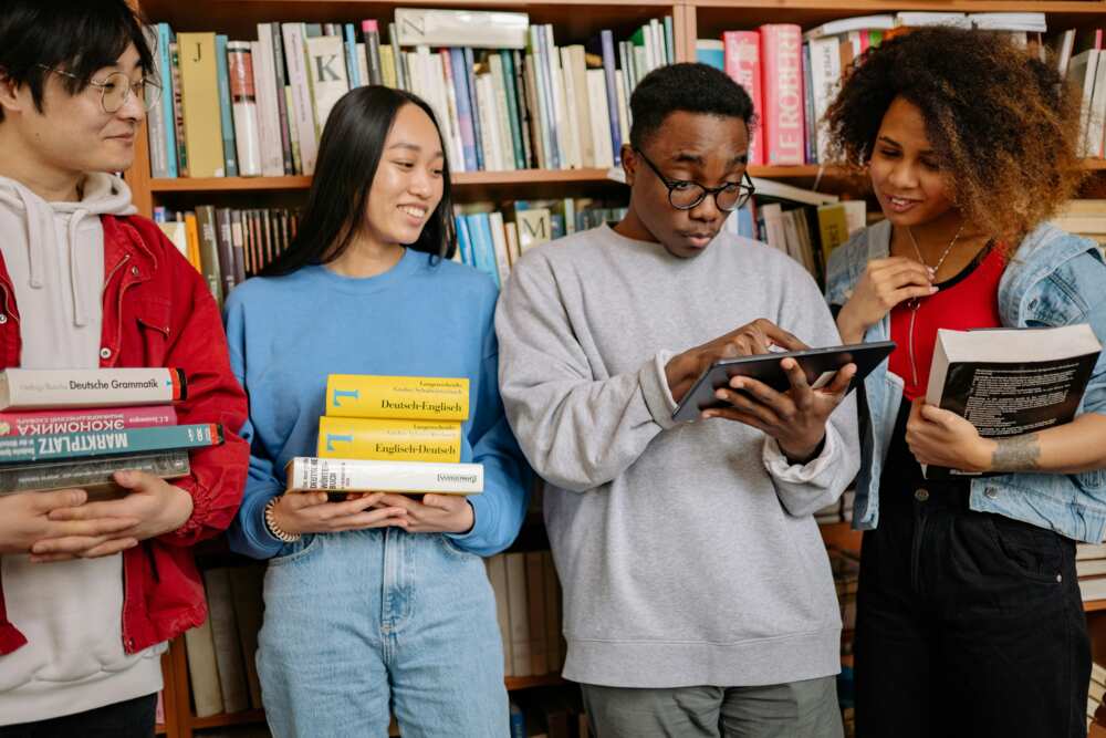 A group of students holding books looking at the digital tablet in a library