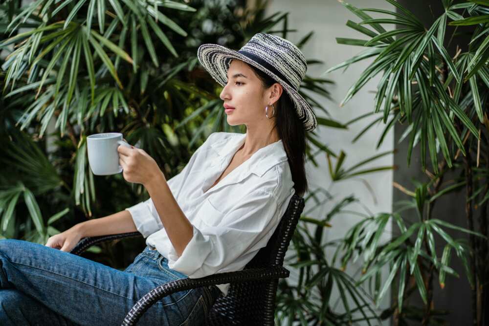 A young female in blue jeans and a white shirt holding hot beverage while resting in an armchair