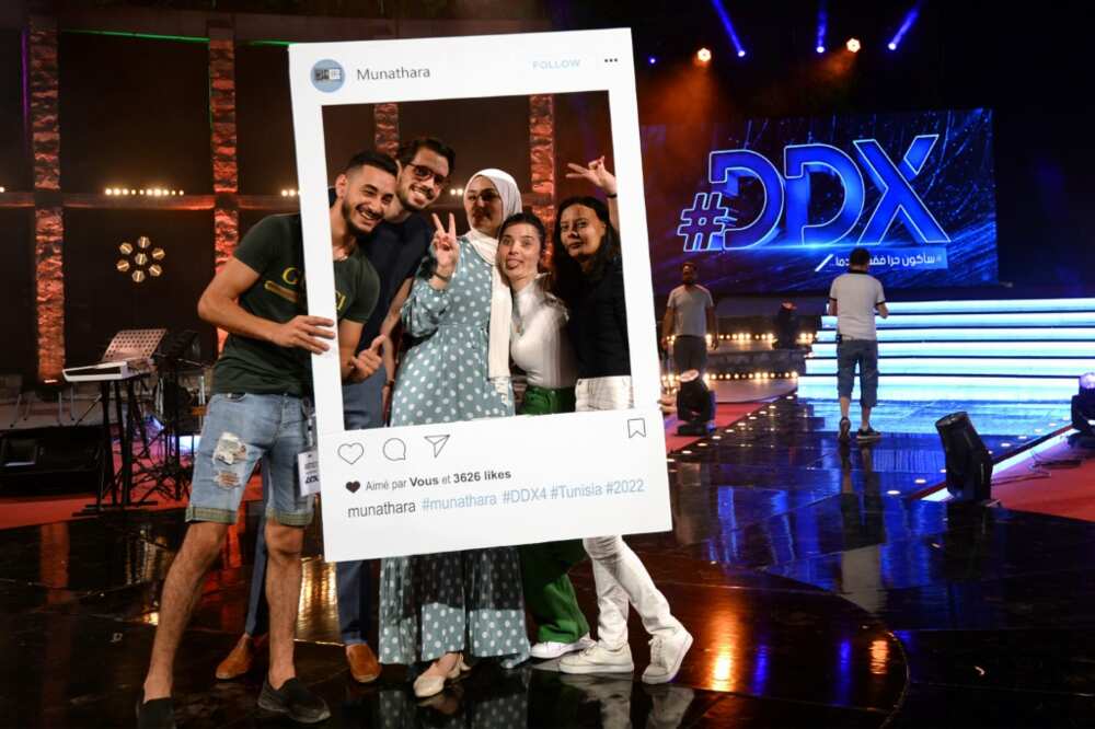 Participants pose for a photo with a cutout frame depicting an Instagram social media post on the set of the #DDX talent show